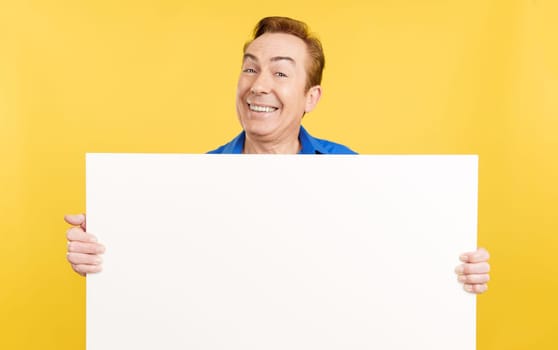 Smiley mature man holding a blank panel in studio with yellow background