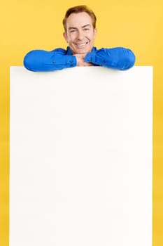 Relaxed mature man leaning on a blank panel in studio with yellow background