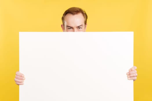 Caucasian mature man hiding behind a blank panel in studio with yellow background
