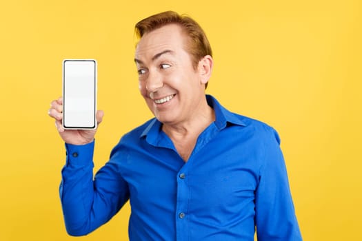 Smiley mature man holding a mobile with a blank screen in studio with yellow background