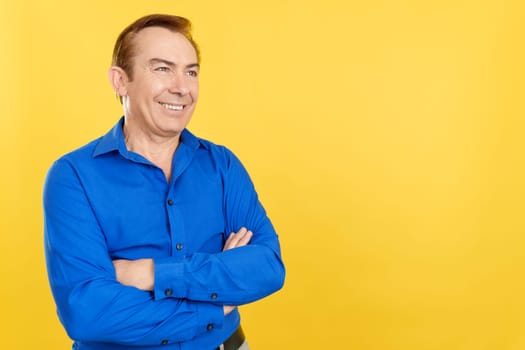 Distracted and smiley mature man standing with arms crossed in studio with yellow background