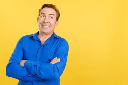 Happy mature man looking aside with arms crossed in studio with yellow background