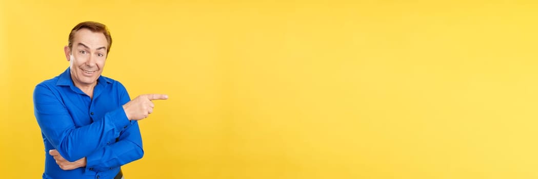 Happy mature caucasian man pointing to the side in studio with yellow background