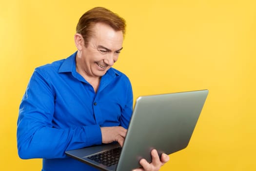 A happy caucasian mature man standing and using a laptop in studio with yellow background