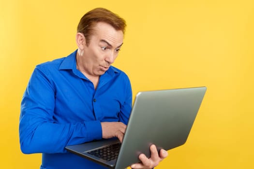 Mature man using a laptop with a surprised look in studio with yellow background