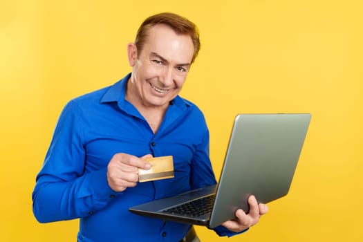 Mature man looking at camera while using a laptop and card to shopping online in studio with yellow background