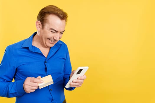 Happy mature man using the mobile and card to shopping online in studio with yellow background