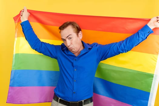 Happy mature man blowing a kiss raising a lgbt rainbow flag in studio with yellow background