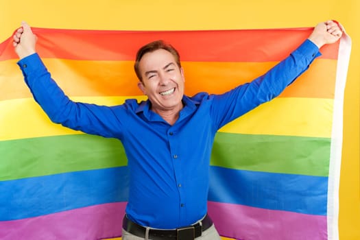 Happy mature man raising a lgbt rainbow flag in studio with yellow background