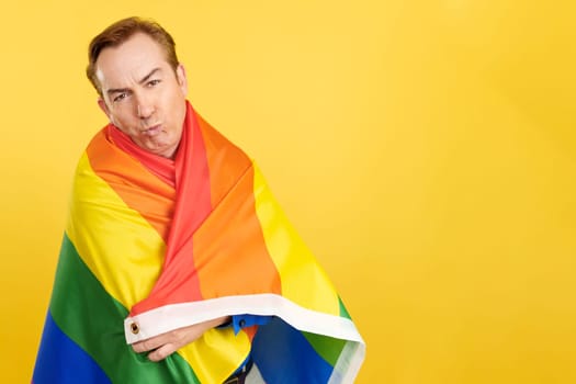 Mature man blowing a kiss while wrapping with a lgbt flag in studio with yellow background