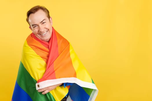 Happy mature man wrapped with a lgbt flag in studio with yellow background