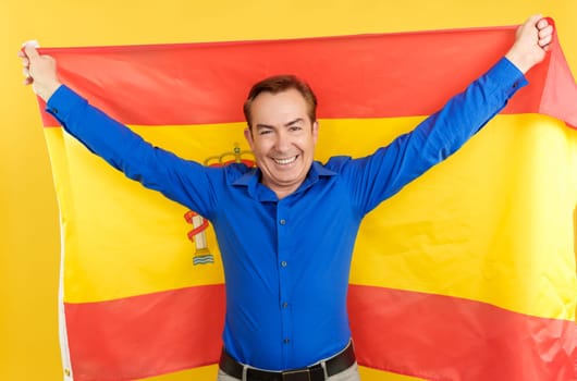 Smiling mature man raising a spanish flag in studio with yellow background