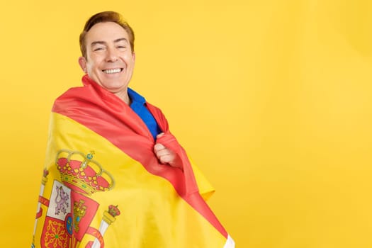 Smiling mature man wrapping with a spanish flag in studio with yellow background