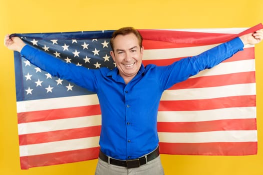Smiley and happy mature caucasian man raising a north america national flag in studio with yellow background