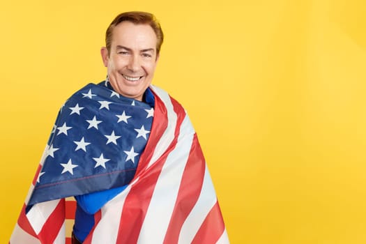 Happy caucasian mature man wrapping with a north america national flag in studio with yellow background
