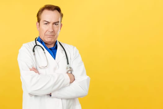 Serious caucasian doctor looking at the camera with arms crossed in studio with yellow background