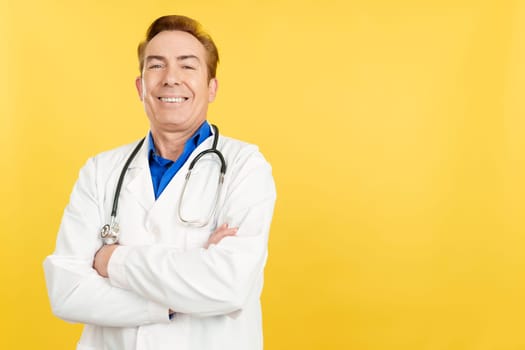 Smiley and mature mature doctor looking at the camera with arms crossed in studio with yellow background