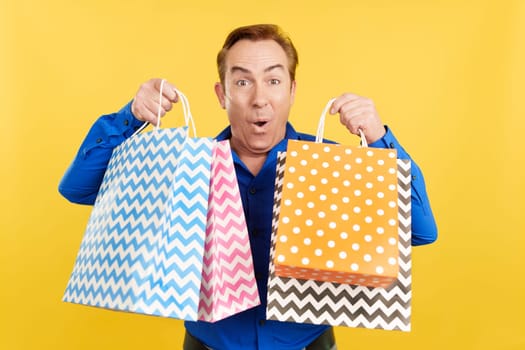 Happy mature caucasian man looking at the camera while showing shopping bags in studio with yellow background