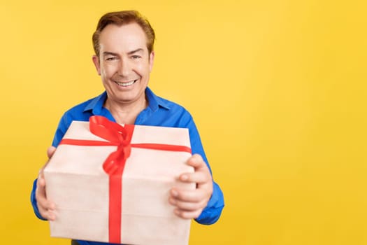 Mature man looking at camera happily holding a present in studio with yellow background