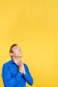 Mature man looking up while praying with folded hands in studio with yellow background