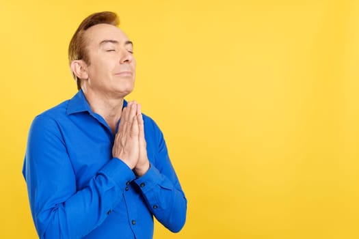 Mature man praying with folded hands and closed eyes in studio with yellow background
