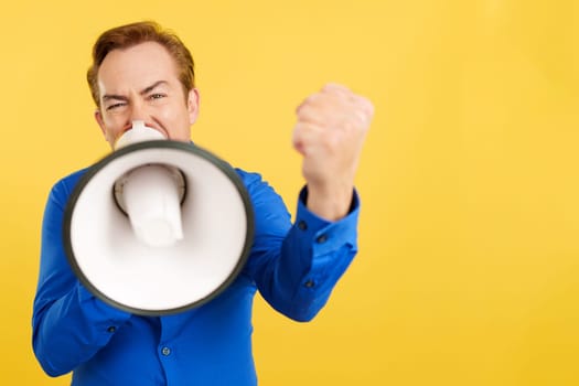 Angry mature man shouting using a loudspeaker in studio with yellow background