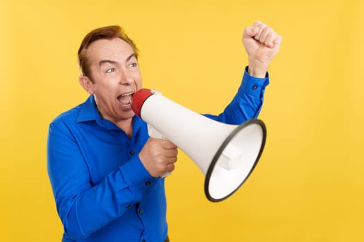Upset mature man shouting using a loudspeaker in studio with yellow background