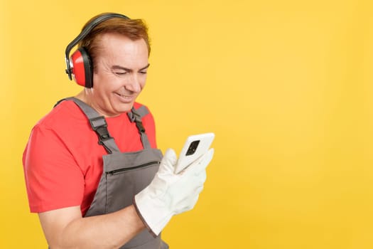 Mature worker in uniform and protective gear using a mobile in studio with yellow background