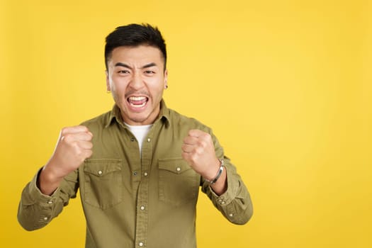 Excited chinese man celebrating while raising arms and fists in studio with yellow background