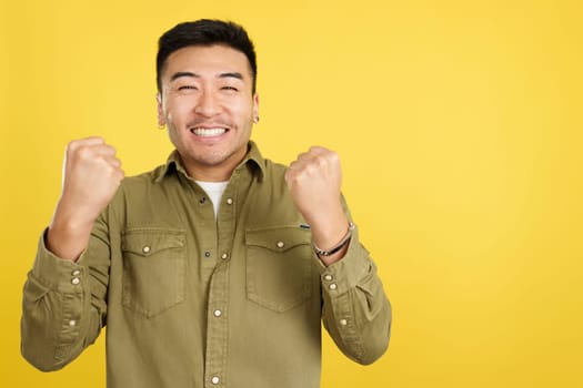 Happy chinese man celebrating while raising fists in studio with yellow background