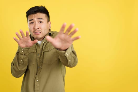 Chinese man gesturing with the hands in fear in studio with yellow background