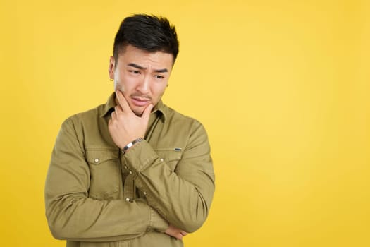 Worried chinese man looking down with pensive expression in studio with yellow background