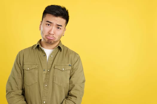Sad handsome chinese man looking at camera in studio with yellow background