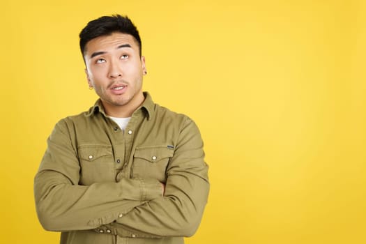 Bored chinese man looking up standing with arms crossed in studio with yellow background