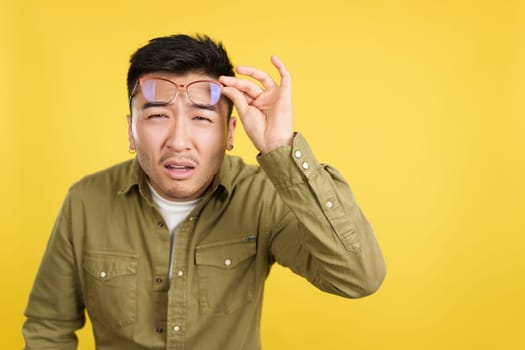 Chinese man taking off her glasses to see while looking at camera in studio with yellow background