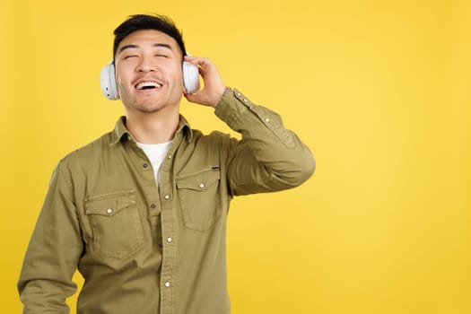 Relaxed chinese man listening to music with headphones connected to a mobile in studio with yellow background