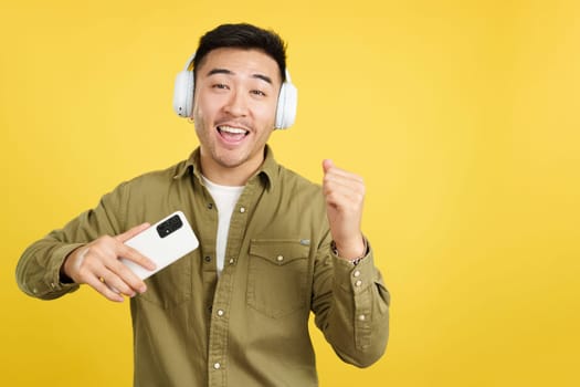 Happy chinese man dancing and listening to music with the mobile phone and headphones in studio with yellow background