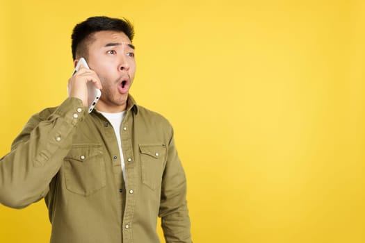 Surprised chinese man opening the mouth while talking to the mobile in studio with yellow background