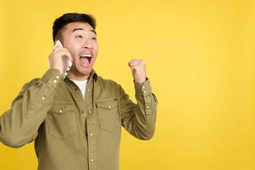 Happy chinese man celebrating while taking to the mobile in studio with yellow background