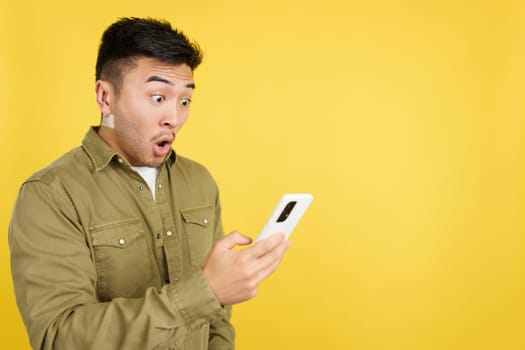 Surprised chinese man looking the screen of a mobile in studio with yellow background