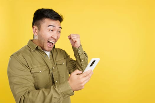 Excited chinese man celebrating while using the mobile phone in studio with yellow background