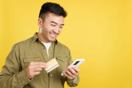 Happy chinese man using the mobile and card to shopping online in studio with yellow background