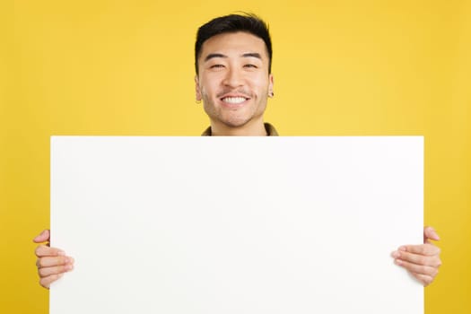 Happy chinese man smiling and holding a blank panel in studio with yellow background
