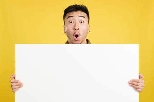 Handsome surprised chinese man hair holding a blank panel in studio with yellow background