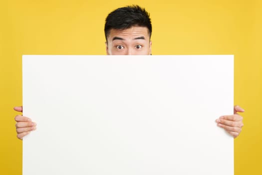 Surprised chinese man hiding behind a blank panel in studio with yellow background