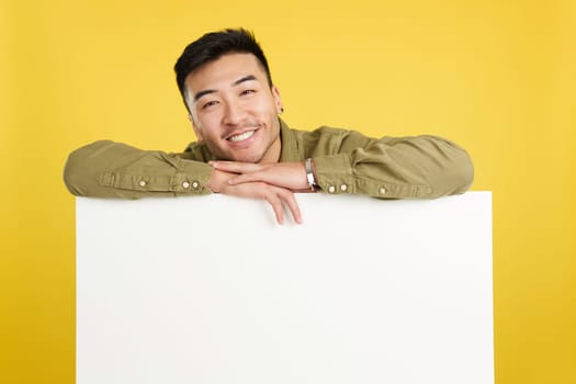 Happy handsome chinese man leaning on a blank panel in studio with yellow background