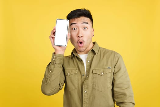 Surprised chinese man holding a mobile with a blank screen in studio with yellow background
