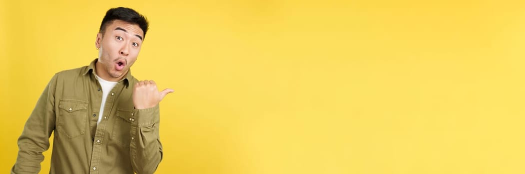 Amazed chinese man pointing surprised to the side in studio with yellow background