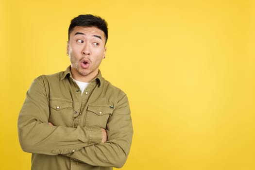 Surprised chinese man standing with open mouth and arms crossed in studio with yellow background