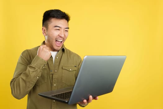 Chinese man celebrating raising the fist while looking at the laptop in studio with yellow background